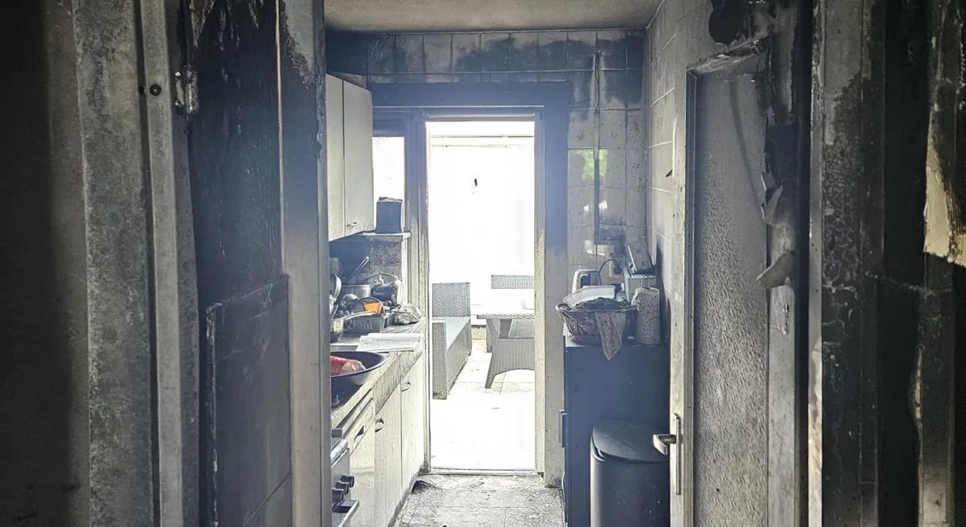 Gang na een woningbrand met zwartgeblakerde wanden en deuren, met de keuken erachter